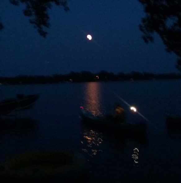 Very blurry photo of a man in a canoe with running lights and a full moon in the background